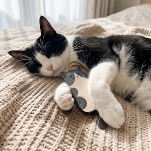Peut inclure: Un porte-clés en cuir plat en forme de chat avec un centre couleur crème, des taches gris foncé, des bords cousus et une petite boucle en cuir marron sur le dessus. Le motif comprend un visage imprimé simple avec des moustaches et un petit nez.