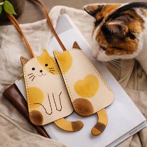 Peut inclure: Deux pochettes en cuir de couleur claire en forme de chats calico avec des taches brunes et jaunes. Chaque pochette présente des détails cousus, un long cordon en cuir fin pour le transport et une queue incurvée au bas de la forme.