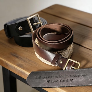 May include: Two leather belts with brass buckles on a wooden surface. One is black, the other brown. A dark grey tag with the text "Self-love isn't selfish. It's survival gear. Love, Sarah" is also visible.