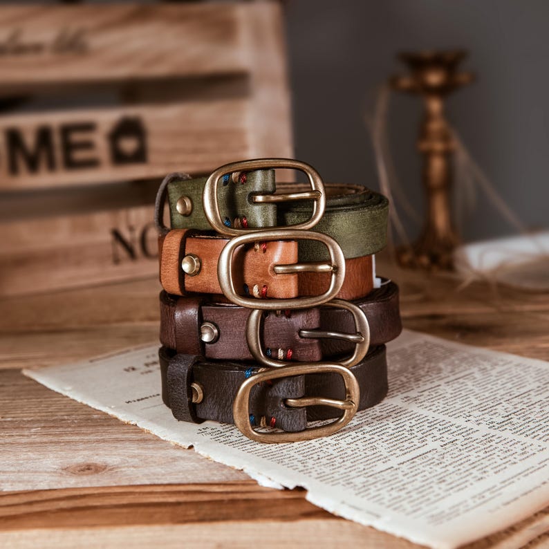 May include: A stack of four leather belts in shades of brown and green, each with a brass buckle. The belts are arranged on a piece of paper with text, suggesting a vintage or rustic aesthetic. The belts vary in color and texture.