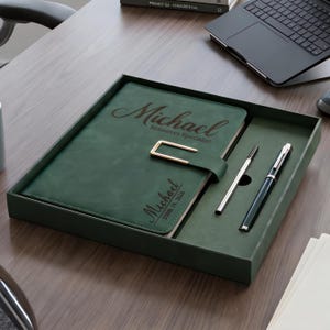 May include: A dark green leather-like notebook set in a matching tray, featuring a gold-tone metal clasp. The cover is engraved with the name Michael, the title Resources Specialist, and the date June 14, 2026. Two pens are included in the tray.