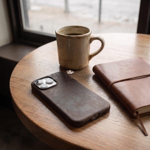 May include: A dark brown leather smartphone case with a triple-lens camera cutout, positioned next to a beige ceramic mug filled with dark liquid and a brown leather-bound notebook on a round wooden surface.
