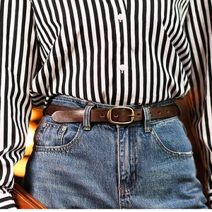 May include: A brown leather belt with a gold-colored buckle is worn with blue denim jeans. The person is wearing a black and white striped button-down shirt. The belt is approximately 1.25 inches wide.