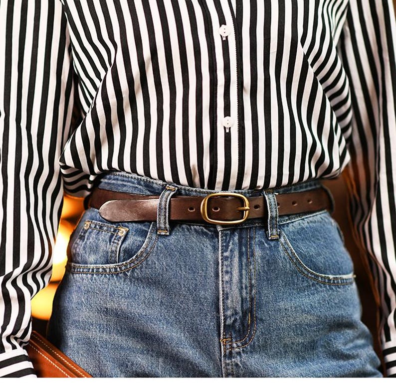May include: A brown leather belt with a gold-colored buckle is worn with blue denim jeans. The person is wearing a black and white striped button-down shirt. The belt is approximately 1.25 inches wide.