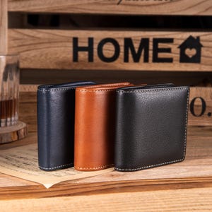 以下が含まれることがあります： ネイビーブルー、ブラウン、ブラックの3つの革製財布。それぞれ白いステッチが施されています。財布は、背景に「HOME」と書かれた木の看板がある木製の表面に表示されています。