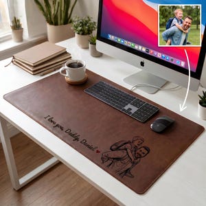 May include: A brown leather desk mat featuring a custom laser-engraved illustration of a child riding on a person's back, accompanied by the text "I love you, Daddy, Daniel" and a small red heart icon, positioned on a white desk surface.