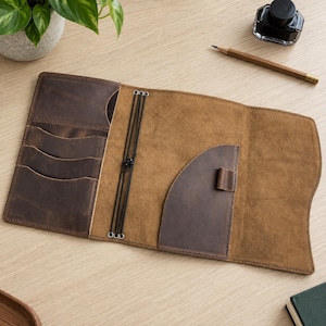 May include: A brown leather notebook cover featuring a tri-fold design with multiple interior card slots, a pen loop, and black elastic bands for securing inserts. The interior displays a smooth tan finish, while the exterior pockets are dark brown.