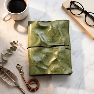 May include: A green leather journal with a black elastic strap. The journal is on a marble surface, alongside a coffee cup, spectacles, and a decorative pen.