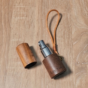 May include: A brown leather Lily Tide perfume atomizer with a silver spray nozzle and a wooden cap. The atomizer has a leather loop and a small gold-colored ring. The wooden cap is next to the atomizer on a wooden surface.