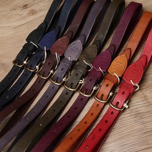 May include: A selection of leather belts in shades of black, navy, purple, green, orange, and red. Each belt has a gold-coloured buckle and a leather tag with text. The belts are displayed diagonally on a wooden surface.