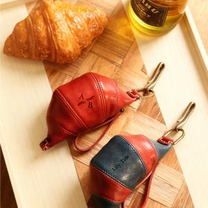 May include: Two leather croissant-shaped keychains, one red and one navy and red, with metal clasps. The red keychain has the initials "A B" and a leaf design. The navy and red keychain has the text "Lily Tide". A croissant and a bottle are in the background.