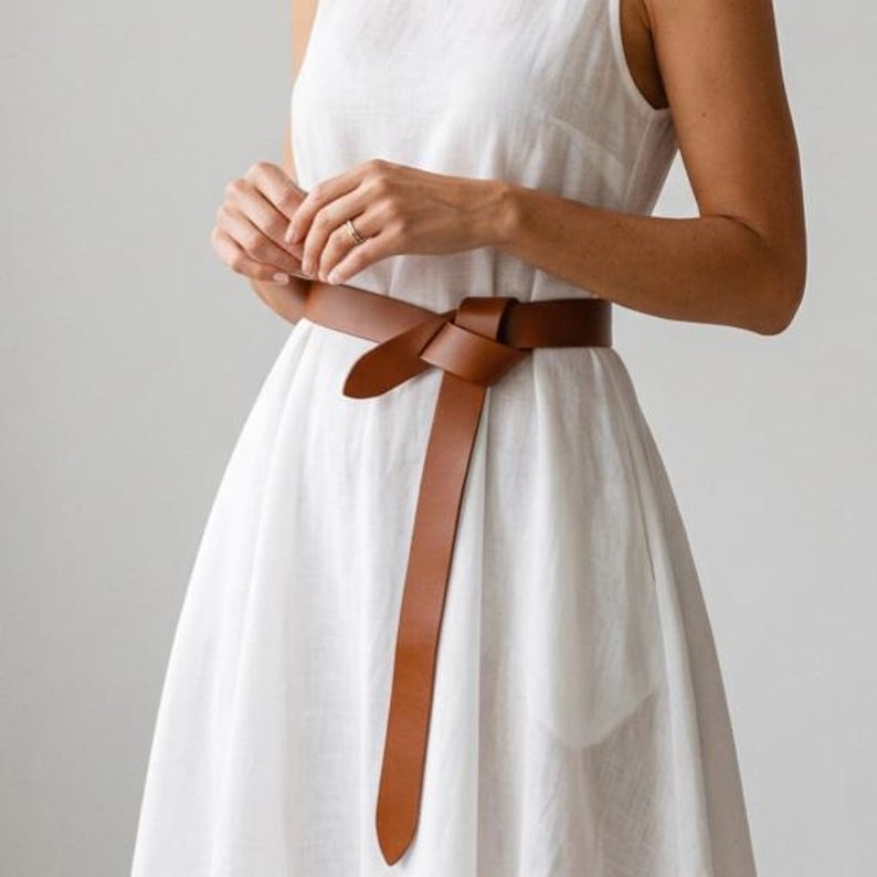 May include: A brown leather belt tied in a knot, worn over a white dress. The belt's long ends hang down, creating a simple yet elegant look. The belt appears to be made of high-quality leather.