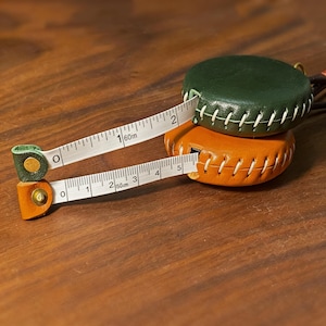 May include: Two leather tape measures, one orange and one green, with white stitching. The tape measures are extended, showing measurements in centimetres and metres. A useful tool for sewing, crafting, and other projects.