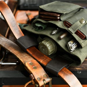 Könnte beinhalten: Nahaufnahme einer olivgrünen Canvas-Tasche mit Lederriemen und verschiedenen Gegenständen. Die Tasche hat mehrere Taschen. Ledergürtel mit Metallbeschlägen sind im Vordergrund. Enthält eine Brieftasche, eine kleine Feldflasche und ein Messer.