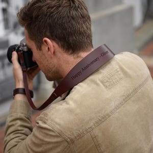 Può includere: Una tracolla per fotocamera in pelle marrone scuro con il testo inciso Happy Anniversary - Oliver. La tracolla è fissata a una fotocamera e presenta una finitura liscia e una struttura resistente progettata per sostenere il peso del dispositivo.
