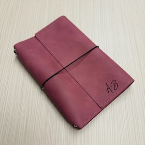Op de afbeelding: Een bordeauxrood leren notitieboek met een klepsluiting en een zwarte elastische band. Het notitieboek heeft de initialen "AB" in de rechter benedenhoek. Het notitieboek is gesloten en ligt plat op een licht houten oppervlak.