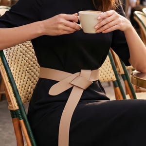 May include: A black dress with a beige belt tied in a knot. The person is holding a beige mug with a brown liquid. The person is sitting in a wicker chair.