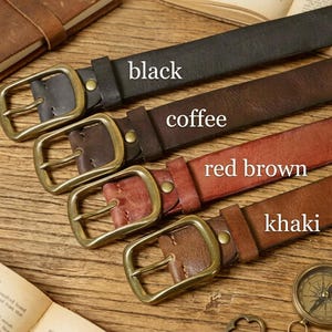 May include: Four leather belts in black, coffee, red brown, and khaki, each with a brass buckle. The belts are displayed on a wooden surface, with the colours labelled. A compass and a journal are visible in the background.