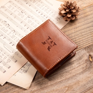 以下が含まれることがあります： 前面に「TAT Martin」のイニシャルがエンボス加工された茶色の革製財布。財布は正方形で、縁に白いステッチが施されています。ヴィンテージの楽譜と木製の表面に置かれています。
