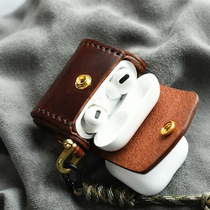 May include: A dark brown leather case for wireless earbuds, featuring a gold-tone metal snap closure and a matching gold-tone metal clip attached to a camouflage-patterned paracord lanyard. The case is open, revealing white wireless earbuds inside.