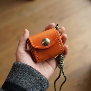 May include: An orange leather wallet with a silver press stud closure. The wallet is held in a hand and has a green and brown braided lanyard. The leather shows visible stitching and a natural grain.