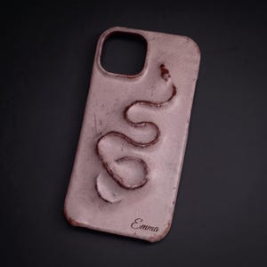 Peut inclure: Une coque de téléphone rose poudré avec un motif de serpent en relief qui serpente verticalement sur la surface. La coque présente une découpe pour l'objectif de l'appareil photo en haut et le nom Emma est imprimé en écriture cursive sombre en bas.