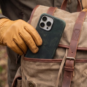 Puede incluir: Una funda de smartphone verde oscuro con recorte para cámara de triple lente, parcialmente insertada en el bolsillo frontal de una mochila de lona color canela con correas de cuero marrón, hebillas de latón y un guante de cuero canela.