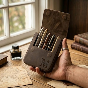 May include: A brown leather pen case with a flap, holding several pens of various colours. The case is embossed with the name "Martin" and a design. The case is held open, revealing the pens inside.