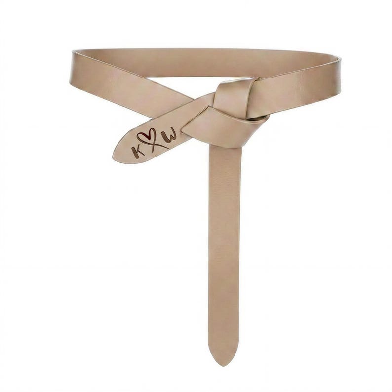 May include: A beige leather belt with a knotted design. The belt features a long, pointed end and a decorative element with the initials "K" and "W" and a heart symbol.