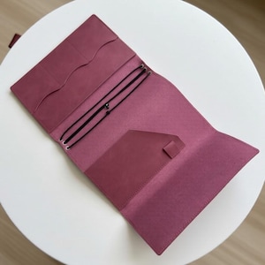 Könnte beinhalten: Ein Notizbuchumschlag aus altrosa Leder mit offenem Innenbereich, ausgestattet mit drei vertikalen Kartenfächern, einer großen Tasche, einer Stiftschlaufe und drei schwarzen Gummibändern zur Befestigung von Heften. Der Umschlag liegt flach.