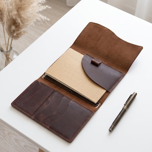 May include: A brown leather notebook cover featuring a fold-over flap, an internal pen loop, and multiple card slots on the left side. The cover holds a tan paper notebook and is accompanied by a matching metallic pen placed to the right side.