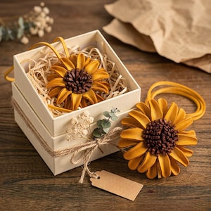 May include: A handcrafted sunflower hair accessory with a brown center and yellow petals. One is displayed in a cream-colored gift box with straw, and another is placed beside the box. The box is tied with twine and a blank tag.