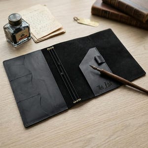 May include: A black leather journal cover featuring three internal elastic bands, a pen loop, and three card slots. The cover includes a dark wooden dip pen resting on the right side, which is embossed with the letter J and a leaf design.