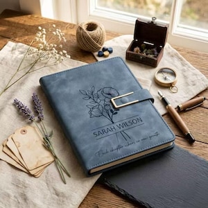 May include: A blue journal with a floral design and the name "SARAH WILSON" printed on the cover. The journal has a gold-colored clasp and the text "Each chapter ushers in new growth". Other items include a compass, pen, and small wooden box.