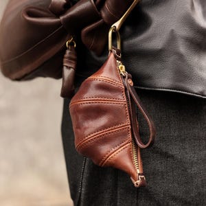 May include: A brown leather pouch with a gold zipper and a wrist strap. The pouch is attached to a larger brown leather bag with a gold-colored clasp. The leather has a textured appearance.