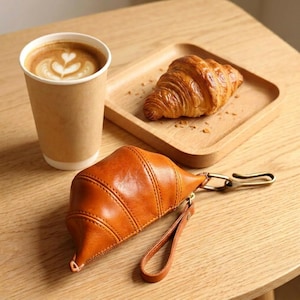May include: A brown leather croissant-shaped pouch with a gold-colored clasp and wrist strap. A paper coffee cup with latte art and a croissant on a wooden tray sit on a wooden table.