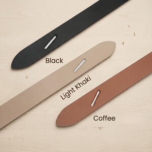 May include: Close-up of three leather straps in black, light khaki, and coffee colors. Each strap has a white stitched detail and is labeled with its respective color name. The straps are arranged diagonally across a light-colored wooden surface.