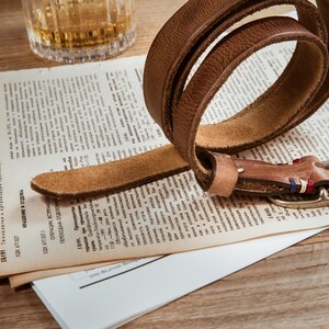 May include: A brown leather belt with a brass buckle is coiled on top of a vintage newspaper. The belt has a textured surface and is partially uncoiled, revealing its full length. A glass of amber liquid sits in the background.