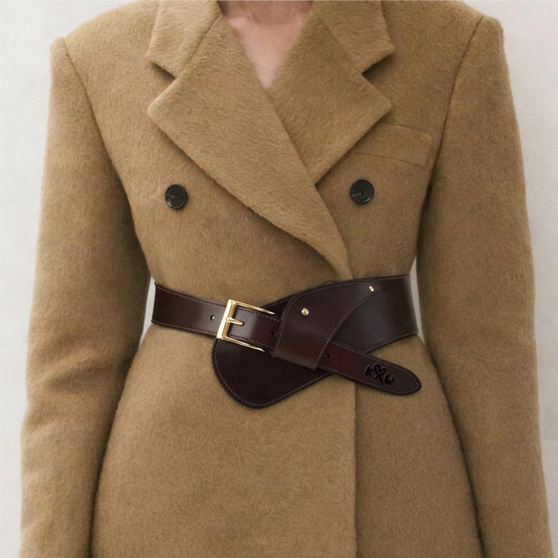 Pu&ograve; includere: Un cappotto doppiopetto beige con una cintura in pelle marrone scuro. La cintura ha una fibbia color oro e un design unico che avvolge la vita. Il cappotto ha due bottoni neri e un classico risvolto.
