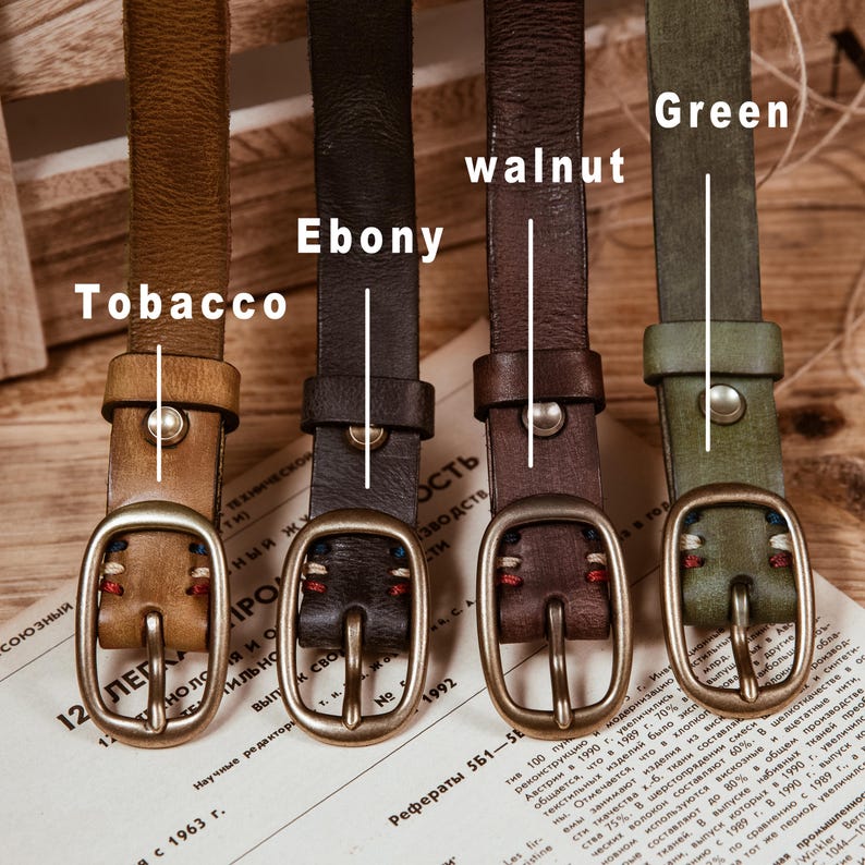 May include: Four leather belts in various colors: tobacco, ebony, walnut, and green. Each belt features a metal buckle and a matching leather loop. The belts are displayed on a printed paper background.