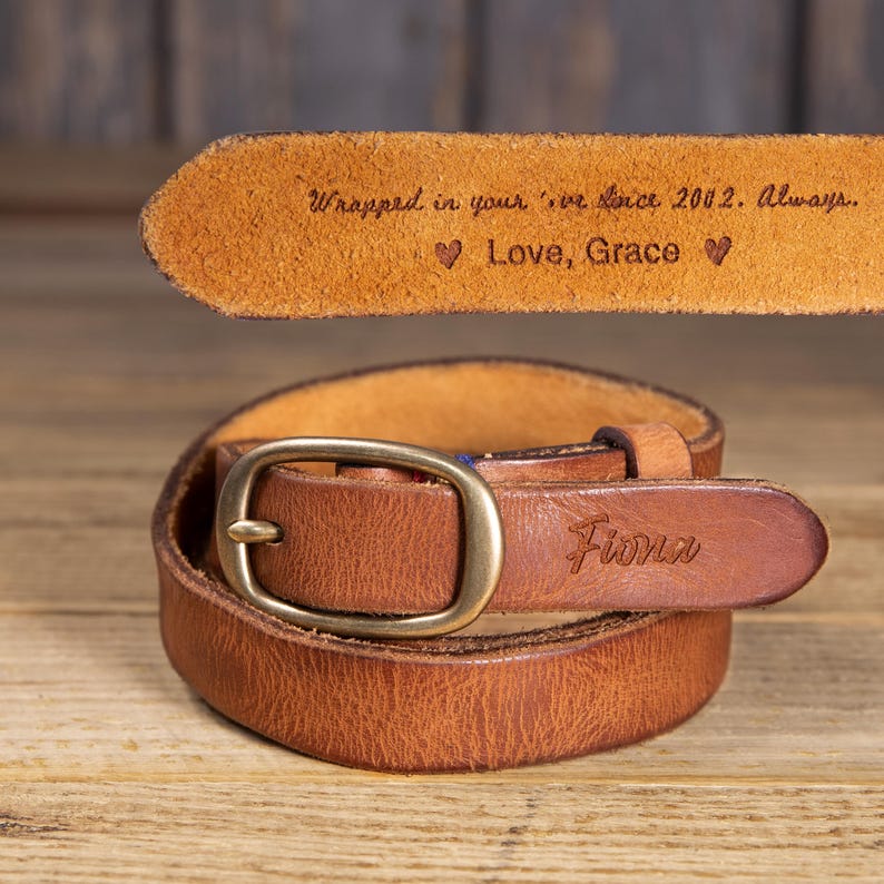 May include: A brown leather belt with a brass buckle. The belt is coiled on a wooden surface. The belt is engraved with the name "Fiona" and the text "Wrapped in your love since 2002. Always... Love, Grace".