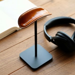 May include: A headphone stand with a brown leather rest and black metal base. The stand is holding a pair of black headphones. The stand is on a wooden surface.