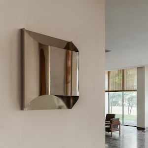 May include: A modern, geometric wall sconce with a polished, reflective metal finish. The sconce has a rectangular shape with curved and angled edges, mounted on a neutral-colored wall. The design is minimalist and contemporary.