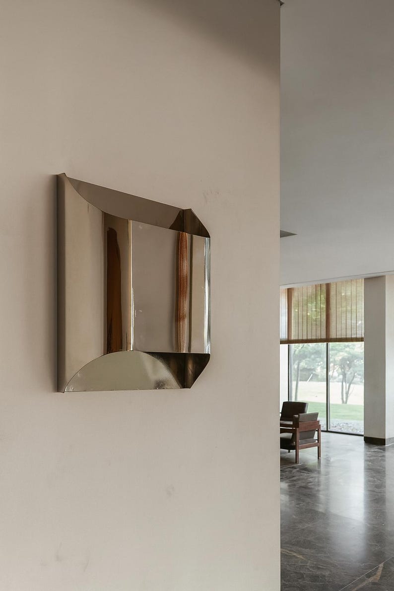 May include: A modern, geometric wall sconce with a polished, reflective metal finish. The sconce has a rectangular shape with curved and angled edges, mounted on a neutral-colored wall. The design is minimalist and contemporary.