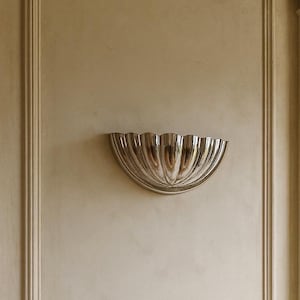 Może przedstawiać: Srebrny kinkiet ścienny w kształcie półokręgu z żłobionym wzorem jest zamontowany na beżowej ścianie. Kinkiet ma odblaskową powierzchnię i jest wyśrodkowany w obramowanej części ściany.