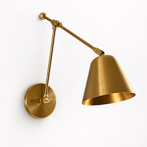 May include: A wall-mounted swing arm lamp with a brushed brass finish. The fixture features a circular wall plate, a long adjustable arm with a central pivot joint, and a conical shade that directs light downwards from the end of the arm.