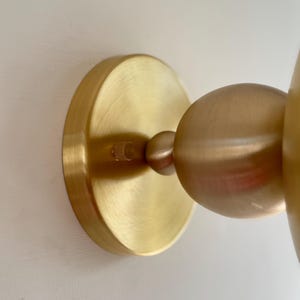 May include: A close-up of a brass wall sconce. The sconce features a circular base and a spherical light fixture. The metal has a warm, golden hue. The sconce is mounted on a white wall.