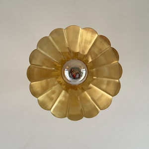 May include: A gold-colored, scalloped-edge ceiling light fixture with a central, clear glass bulb. The fixture has a flower-like design and is mounted on a white ceiling. The light reflects in the bulb.