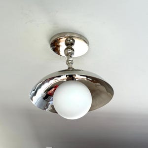 May include: A ceiling-mounted light fixture featuring a polished silver-tone metal canopy and a matching wide, shallow dome-shaped shade. A central stem with spherical accents supports a single, spherical white frosted glass globe light bulb.