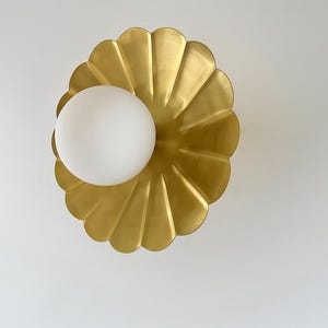 May include: A gold-colored, scalloped wall sconce with a white, spherical light fixture. The sconce has a petal-like design, and the light is positioned in the center. The fixture is mounted on a white wall.